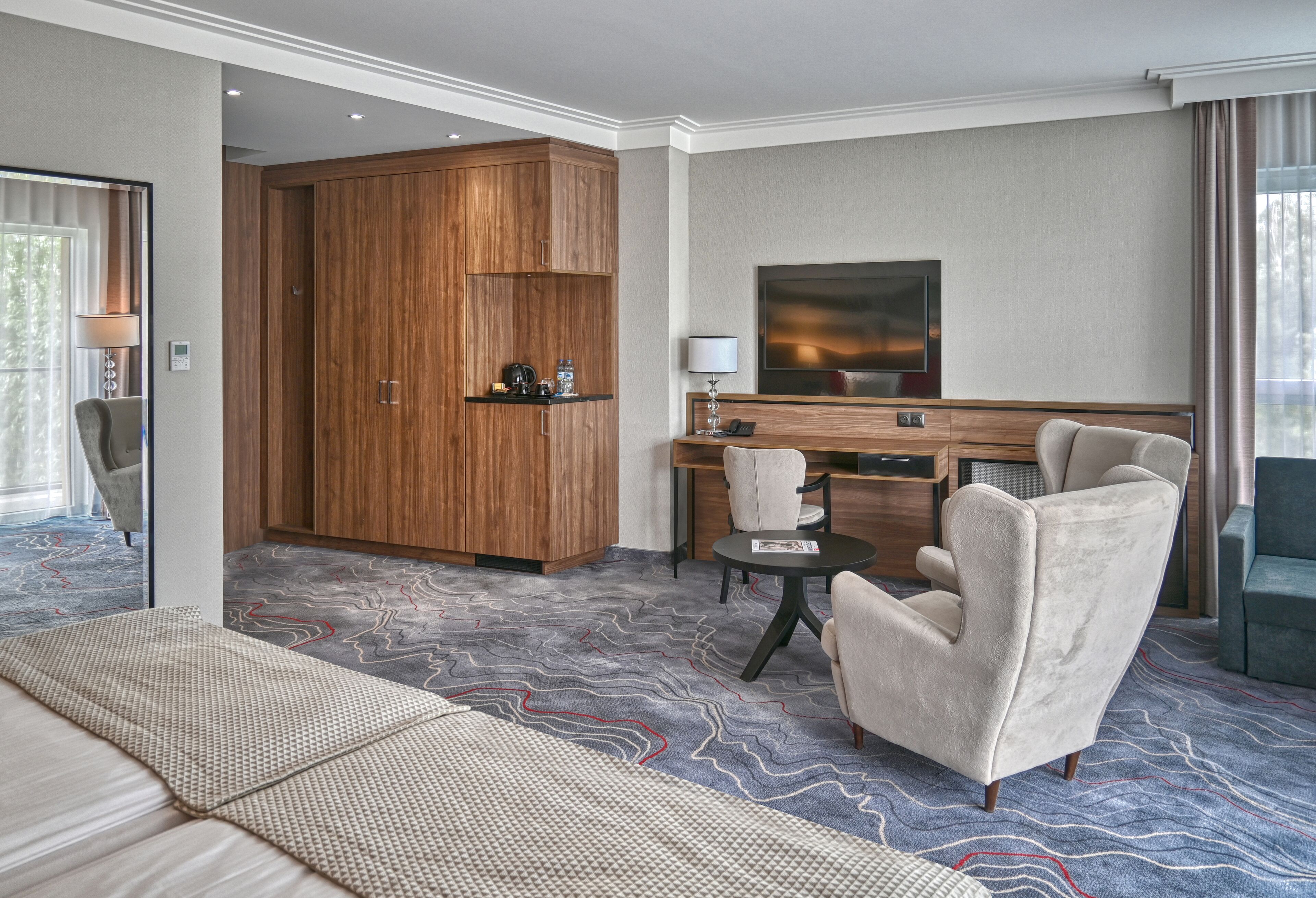 A hotel room with a large wardrobe, a desk with a chair, a television, and a patterned carpet.