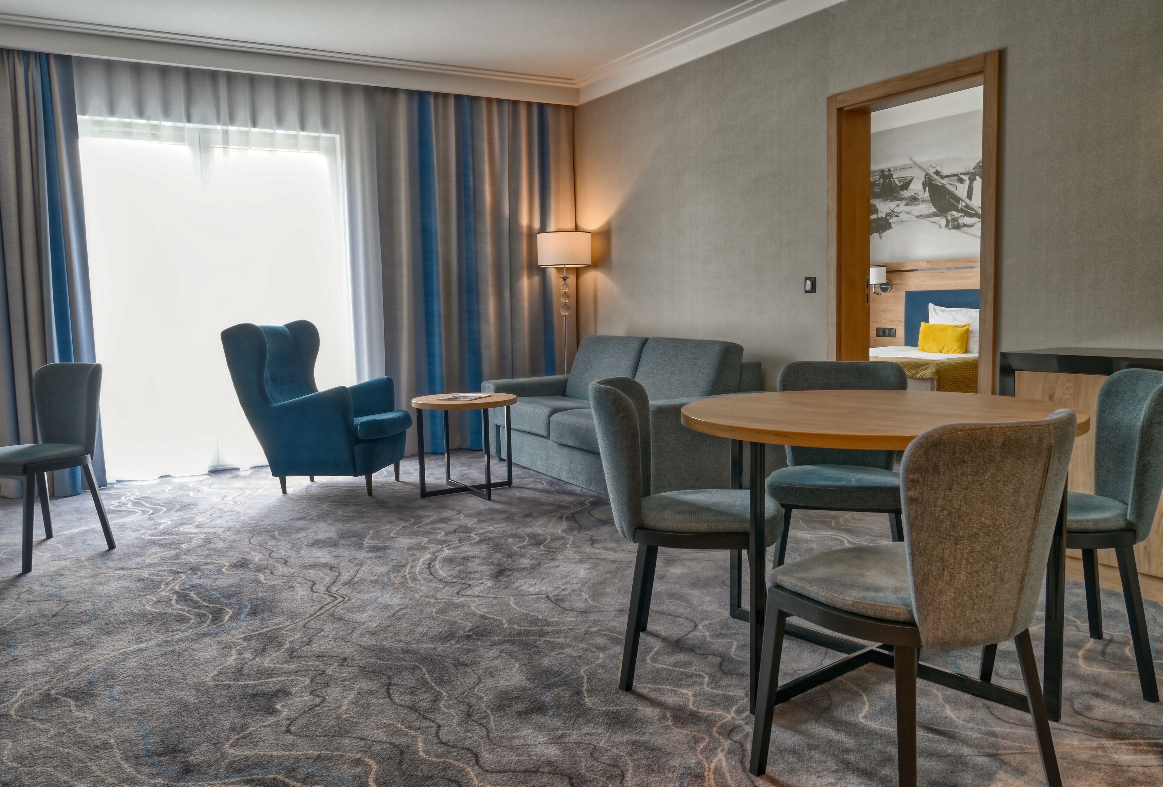 A hotel room with a large window, a sofa, two armchairs, a small table, and a round dining table with chairs.