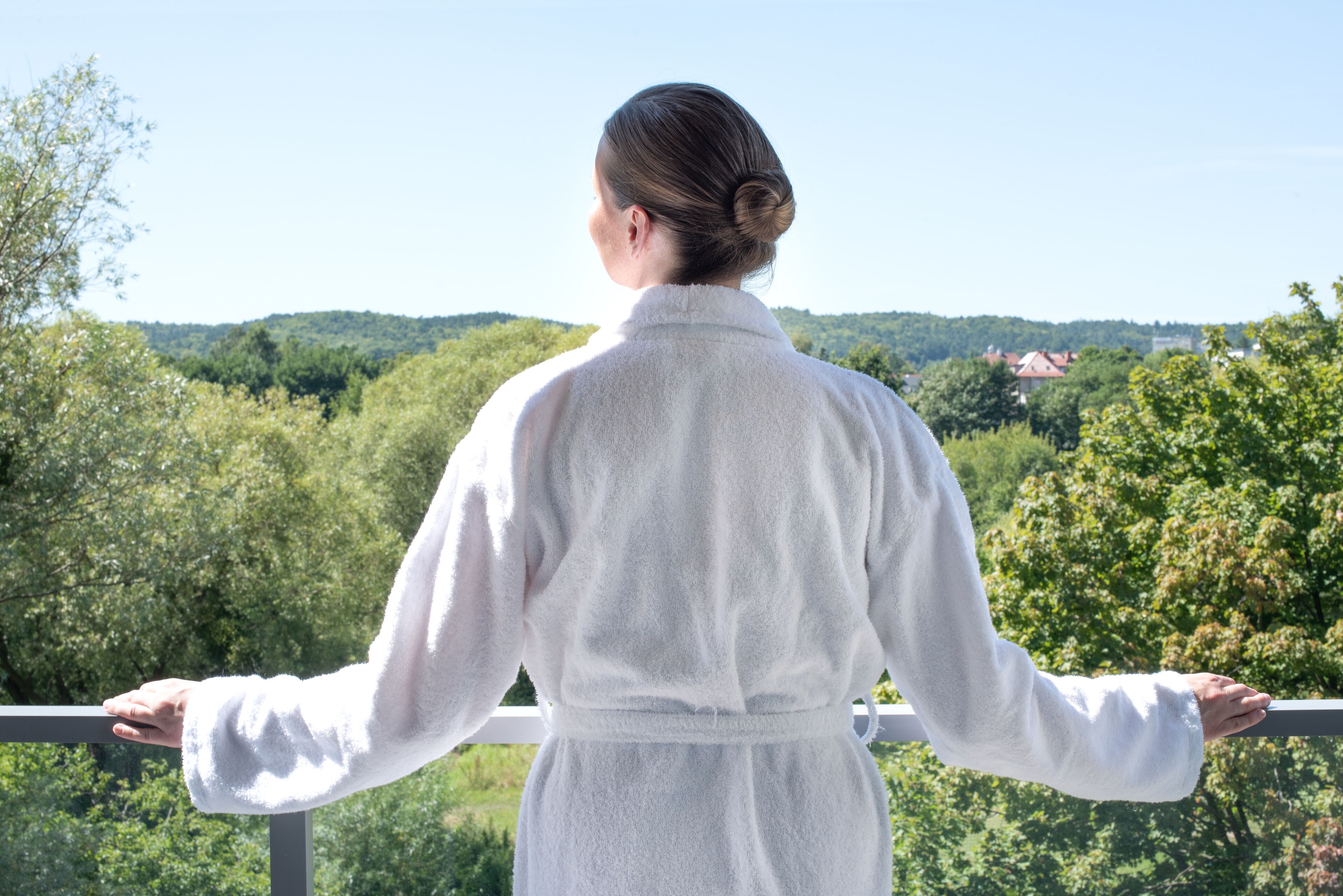A person in a white robe standing on a balcony with a view of a forested area and houses in the distance.