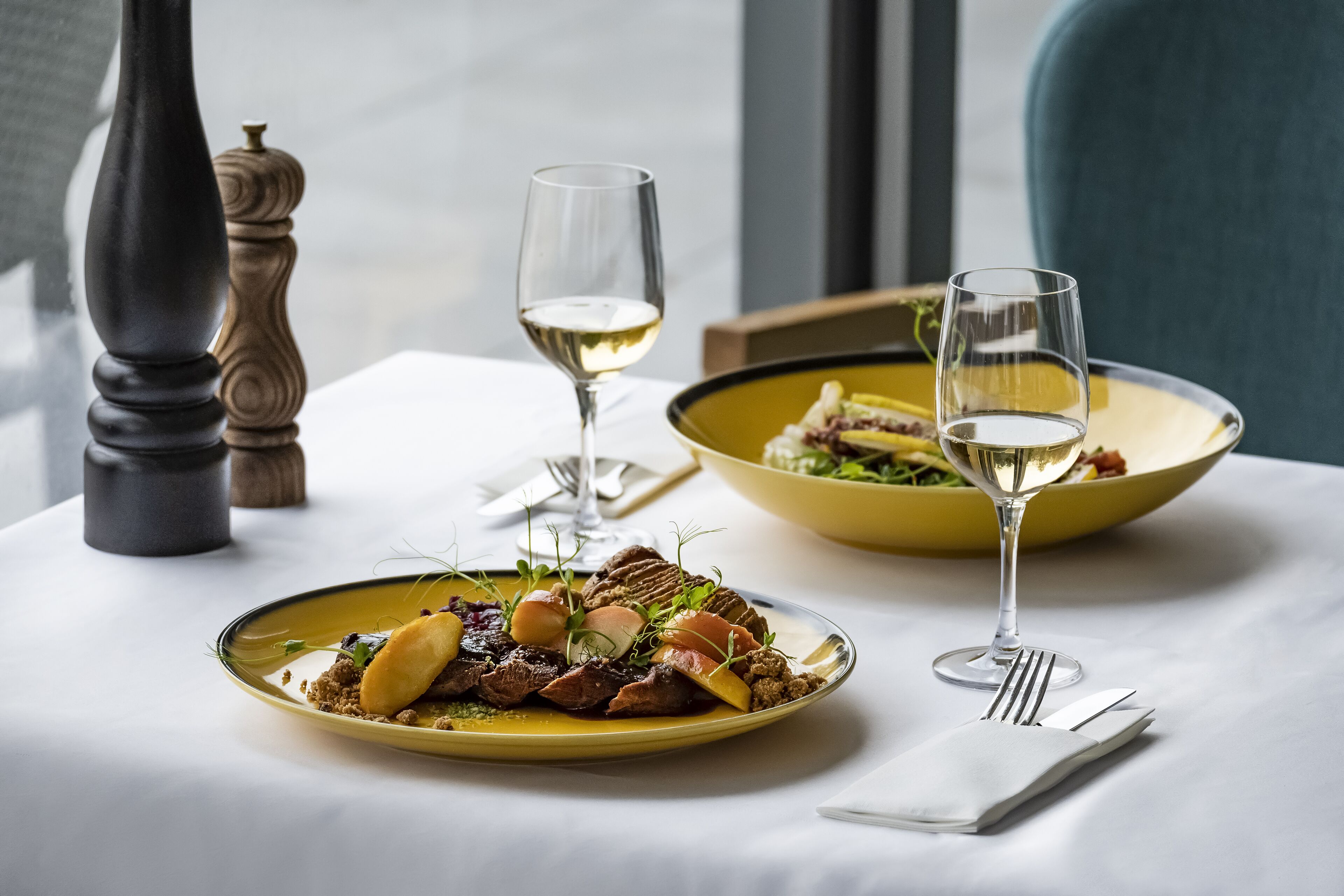 A table with two plates of food, two wine glasses, and a salt and pepper shaker.