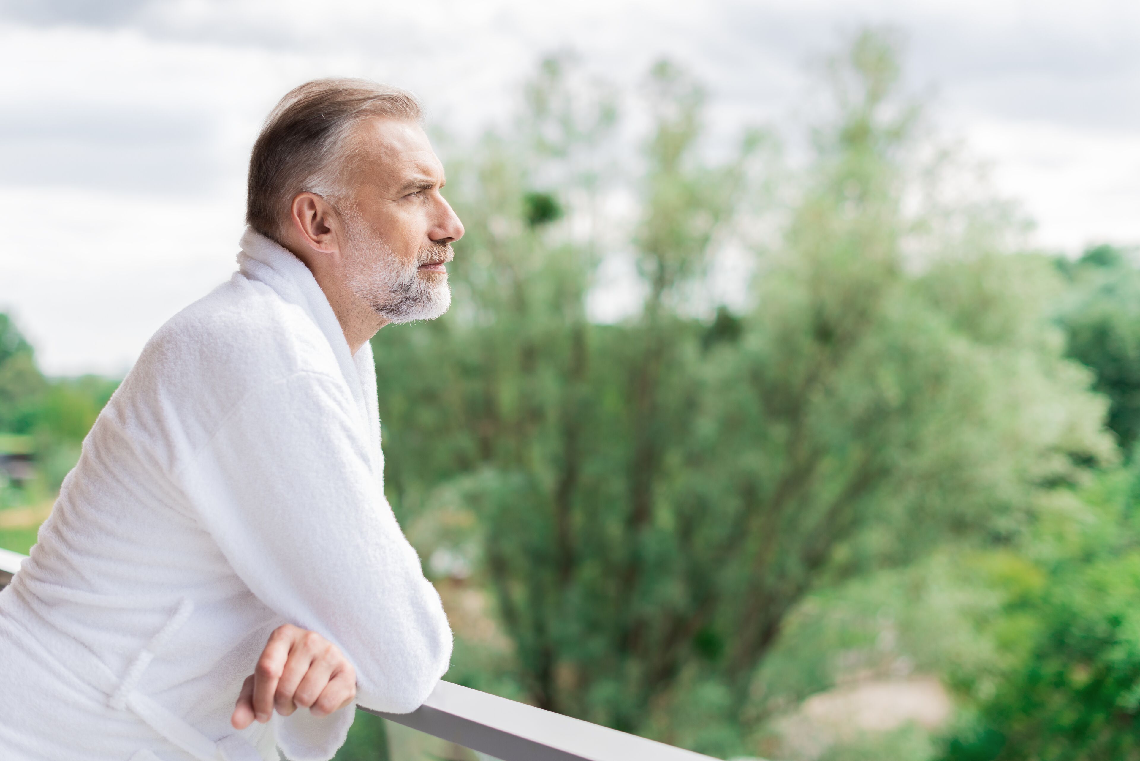 A person leaning on a balcony railing with a view of trees and a cloudy sky.