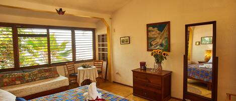 A bedroom with a bed, a dresser, a mirror, a window with blinds, a chair, and a small table with a vase of flowers.