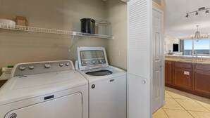 Un espace buanderie équipé d’un lave-linge et d’un sèche-linge à chargement frontal, d’une étagère avec des récipients, et offrant une vue sur la cuisine et l’extérieur.