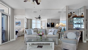 A spacious living room with a large sofa, a coffee table, and a television. There is a dining area with a glass-top table and chairs. The room has large windows and a ceiling fan. - Jade East 1220 (Destin)