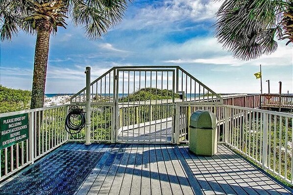 Une terrasse en bois avec une porte en métal, des palmiers et un panneau indiquant qu’il faut rincer les accessoires de plage avant d’entrer.