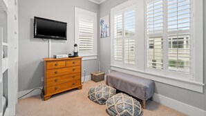 A bedroom with a TV, wooden dresser, ottomans, and large windows with blinds.