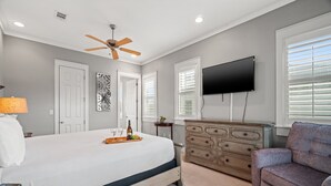 A bedroom with a bed, dresser, TV, ceiling fan, and windows.