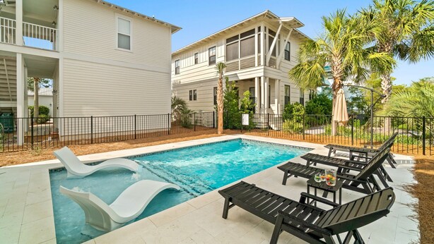 A swimming pool with lounge chairs and a small table, surrounded by a fence and palm trees.