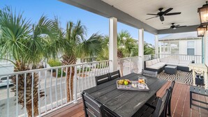 A covered outdoor patio with a dining table, chairs, and a sofa. There are ceiling fans and a lantern. Palm trees are visible in the background.