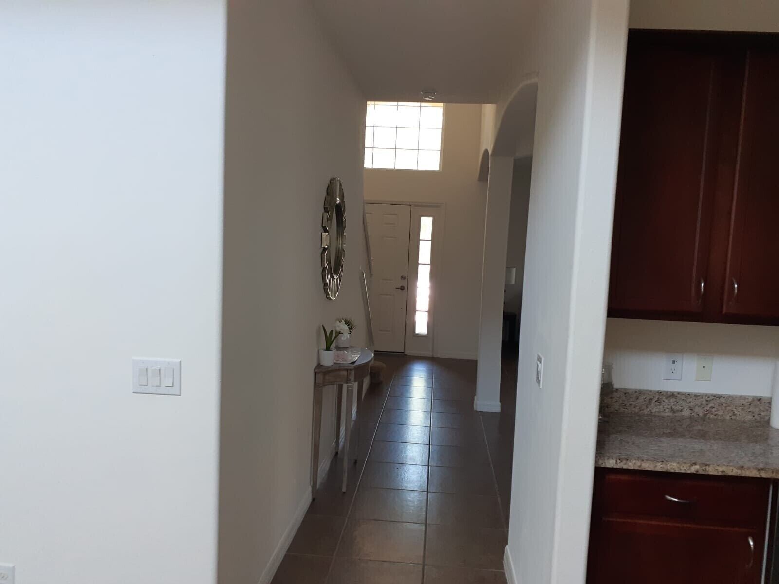 Un couloir étroit, dont le sol est recouvert de carreaux, mène à une porte dotée d’une fenêtre. On y trouve une petite table sur laquelle est posée une plante en pot, ainsi qu’un miroir décoratif accroché au mur.