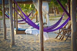 A beach setting with a hammock, wooden chairs, and a table under a thatched roof.