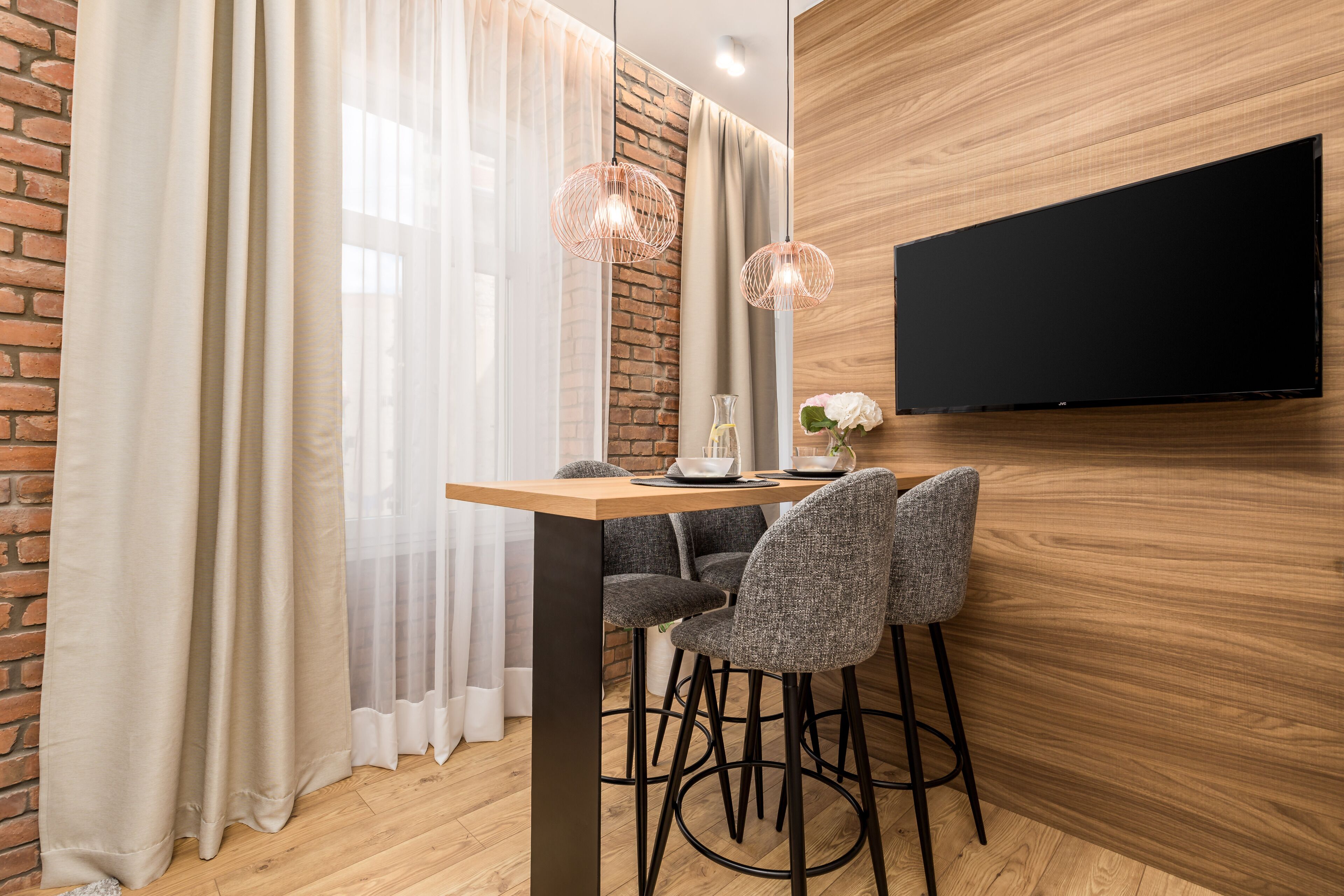 A modern dining area with a wooden table, two grey chairs, a black TV mounted on the wall, and a brick wall.