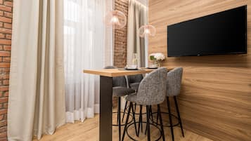 A modern dining area with a wooden table, two grey chairs, a black TV mounted on the wall, and a brick wall.