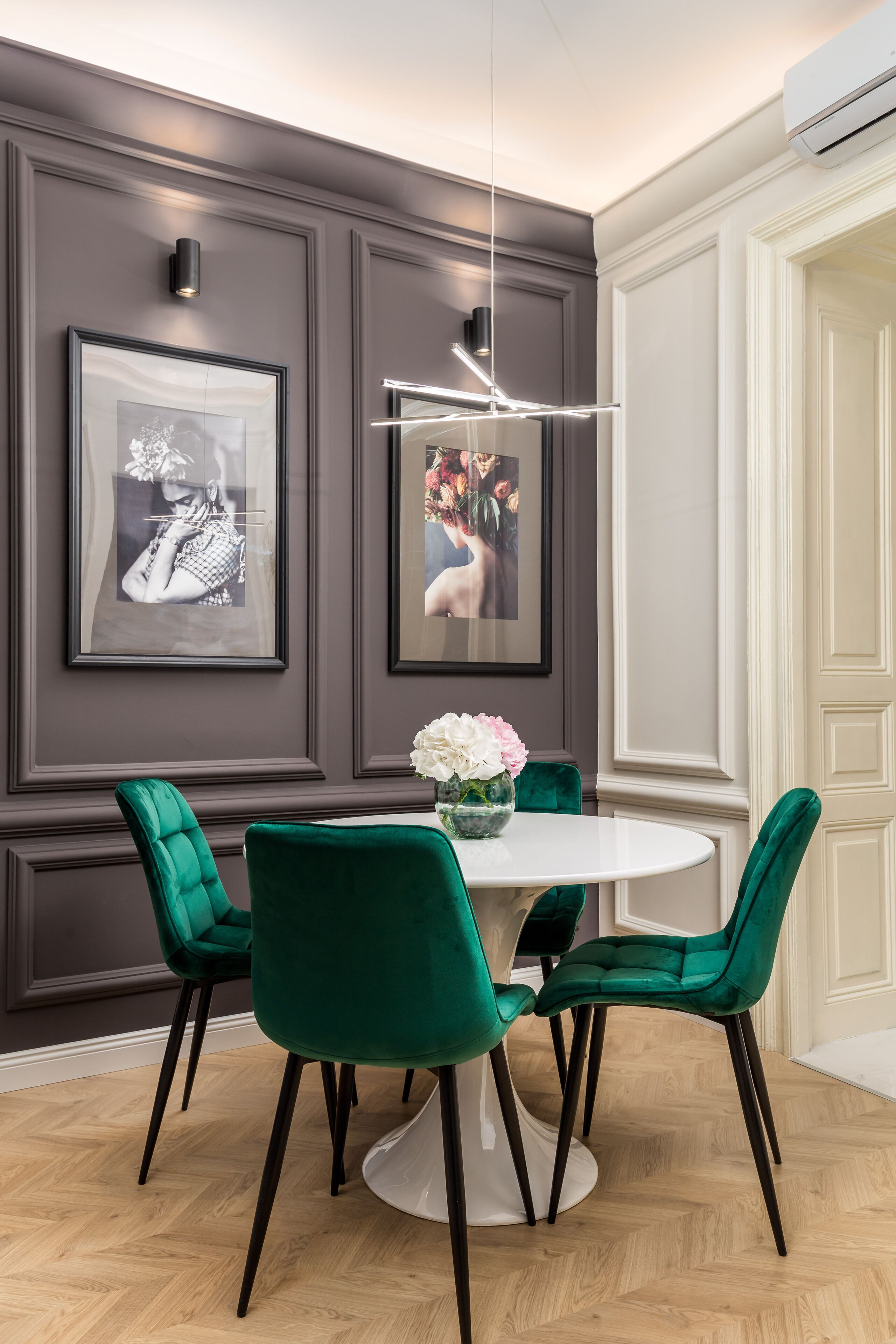 A dining area with a round table, green velvet chairs, a white vase with flowers, and framed pictures on the wall.