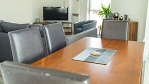 A modern living room with a wooden dining table, grey sofas, and a flat-screen TV.