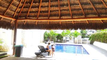 A thatched-roof outdoor area with a pool, lounge chairs, and a trash bin.