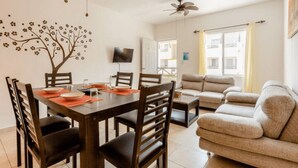 A dining area with a wooden table, chairs, and a wall-mounted TV. There is a sofa and a window with curtains. - Nice Apartment & Pool in Playa del Carmen 8 Min From the Caribbean Sea (Playa del Carmen)