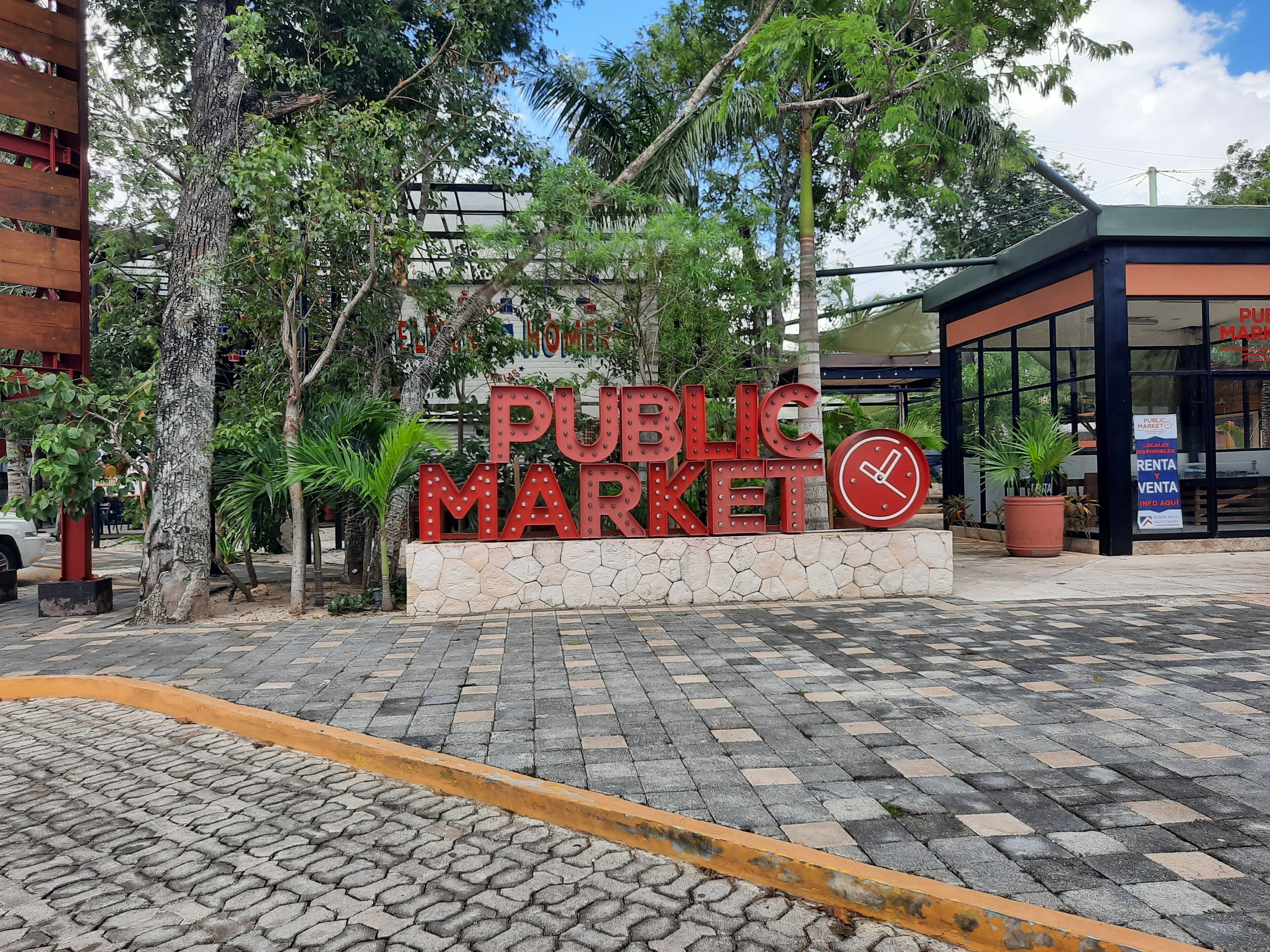 A public market entrance with a sign, trees, and a building in the background.