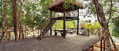 A wooden deck with a thatched-roof structure, surrounded by trees.