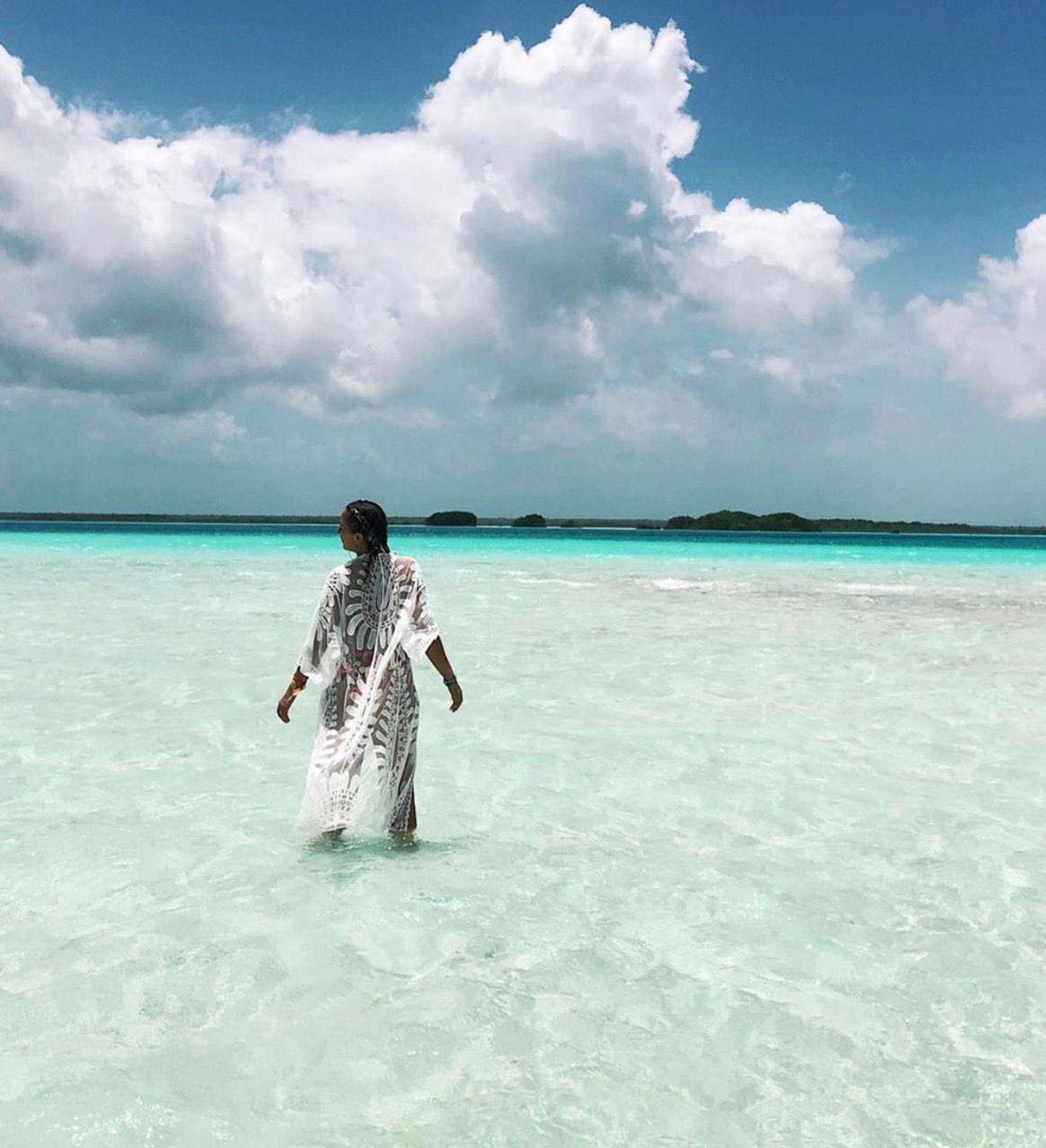 A person walking in clear turquoise water with a distant island and cloudy sky.