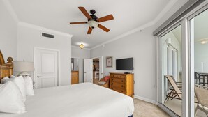 Ein Schlafzimmer mit einem Bett, einem Deckenventilator, einem Fernseher, einem Schminktisch und einer Glasschiebetür, die auf einen Balkon mit Gartenmöbeln führt.