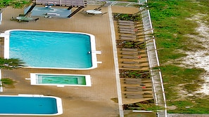 Vista aérea de un complejo turístico con una piscina rectangular grande, una piscina más pequeña y un área de estar cubierta con mesas y sillas.