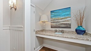Un couloir avec un comptoir en granit, un tableau représentant une scène de plage, un vase contenant des plantes séchées et une lampe fixée au mur.