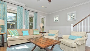 Un salon avec un canapé beige, une table basse en bois et un fauteuil de couleur crème. Des rideaux sont accrochés aux fenêtres, une lampe est posée sur une table d’appoint, et on peut voir le paysage à travers les fenêtres.