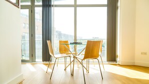 A dining area with a glass table and four chairs, large windows with curtains, and a view of buildings outside. - Kent Street - Apt 4 (Liverpool)