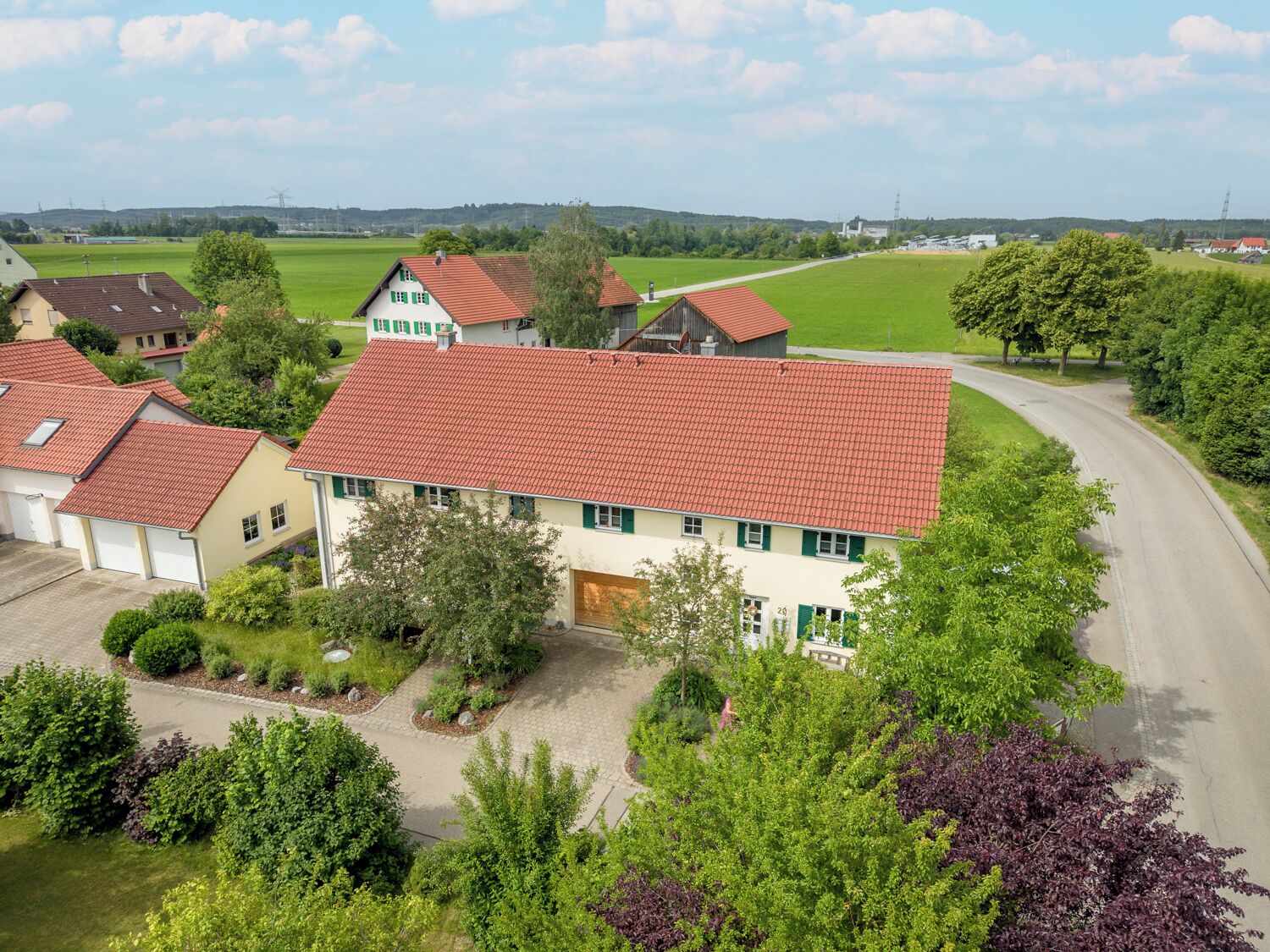 A residential area with houses featuring red-tiled roofs, surrounded by green fields and trees.
