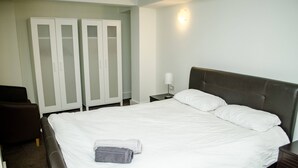 Ein ordentlich bezogenes Bett mit weißen Leinen und zwei Handtüchern, ein dunkelbraunes Kopfstück, ein Nachttisch mit einer Lampe und ein Kleiderschrank mit Milchglas-Türen.