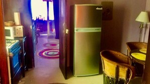 Eine kleine Küche mit Kühlschrank, Mikrowelle und Herd. Auf dem Boden liegt ein farbenfroher Teppich und ein Stuhl ist zu sehen.