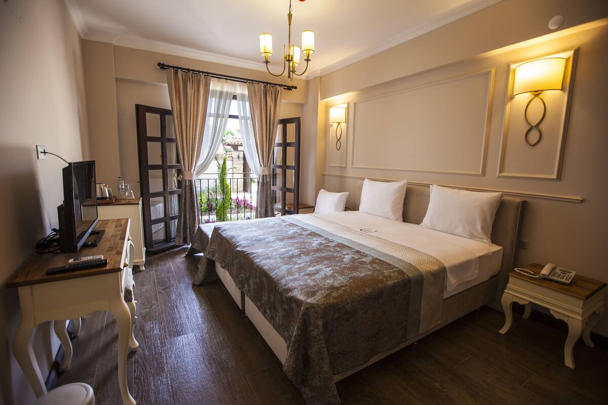 A hotel room with a large bed, bedside tables, a desk with a computer, and a view of a garden through an open door.