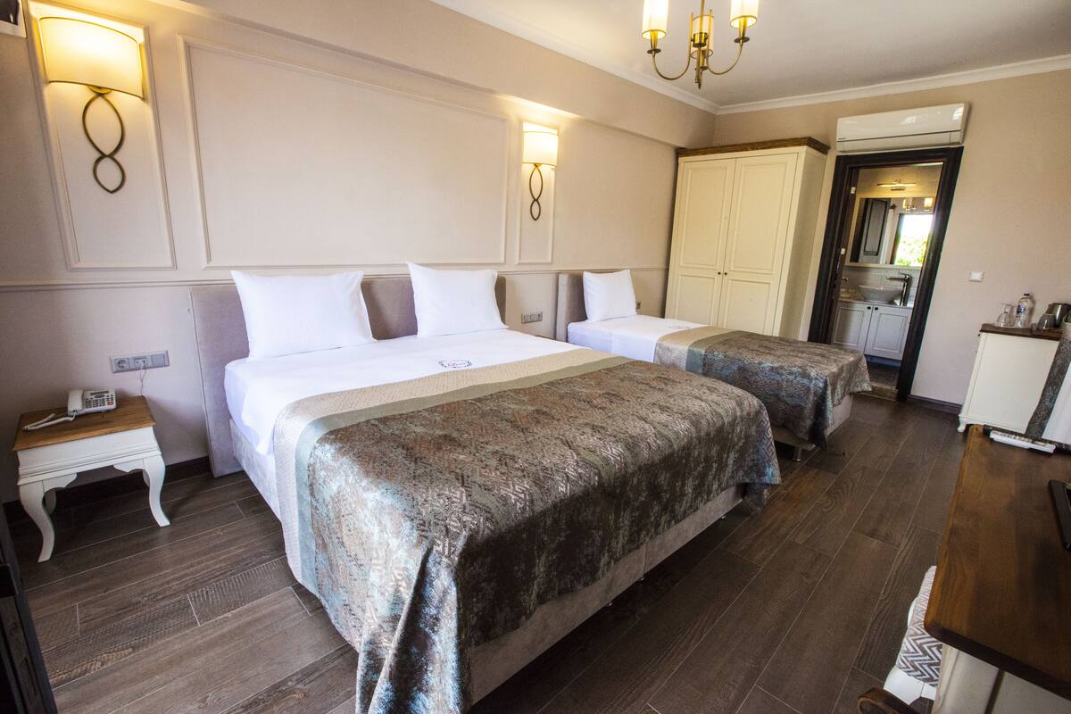 A hotel room with two beds, a small bedside table with a phone, a wardrobe, and a kitchenette area.