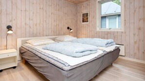 A bedroom with a wooden paneled wall, a single bed with a grey mattress and blue bedding, two bedside lamps, a small white nightstand, and a window with blinds.