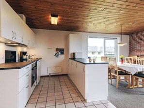 A compact kitchen with white cabinets, a black countertop, and a tiled floor. There is a dining area with wooden chairs and a table, and a brick wall in the background.
