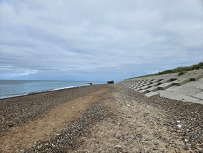 A pebbled beach with a concrete sea wall and a grassy embankment. - 7 Person Holiday Home in Frostrup (Frøstrup)