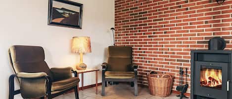 Een gezellige woonkamer met twee fauteuils, een bijzettafeltje met een lamp, een open haard en een ingelijst schilderij aan de muur.