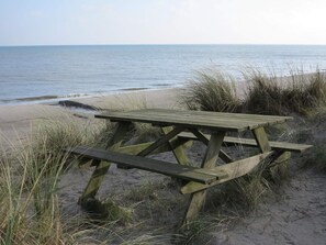 A wooden picnic table on a sandy beach with grass in the foreground and the ocean in the background. - 6 Person Holiday Home in Albaek (Jerup)