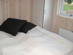 A bed with white bedding and two black pillows, a wooden wall, and a window with a view of greenery.