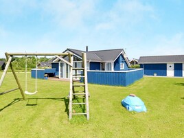 Un parque infantil con un columpio y una escalera de madera, una casa azul con patio cercado y un cielo despejado.