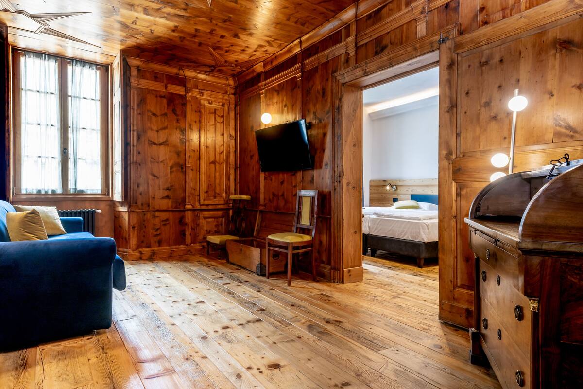 Ein holzgetäfeltes Zimmer im Chalet-Stil mit einem blauen Sofa, einem an der Wand befestigten Fernseher, einem kleinen Tisch mit Stuhl und einem Bett mit weißem Kopfteil.