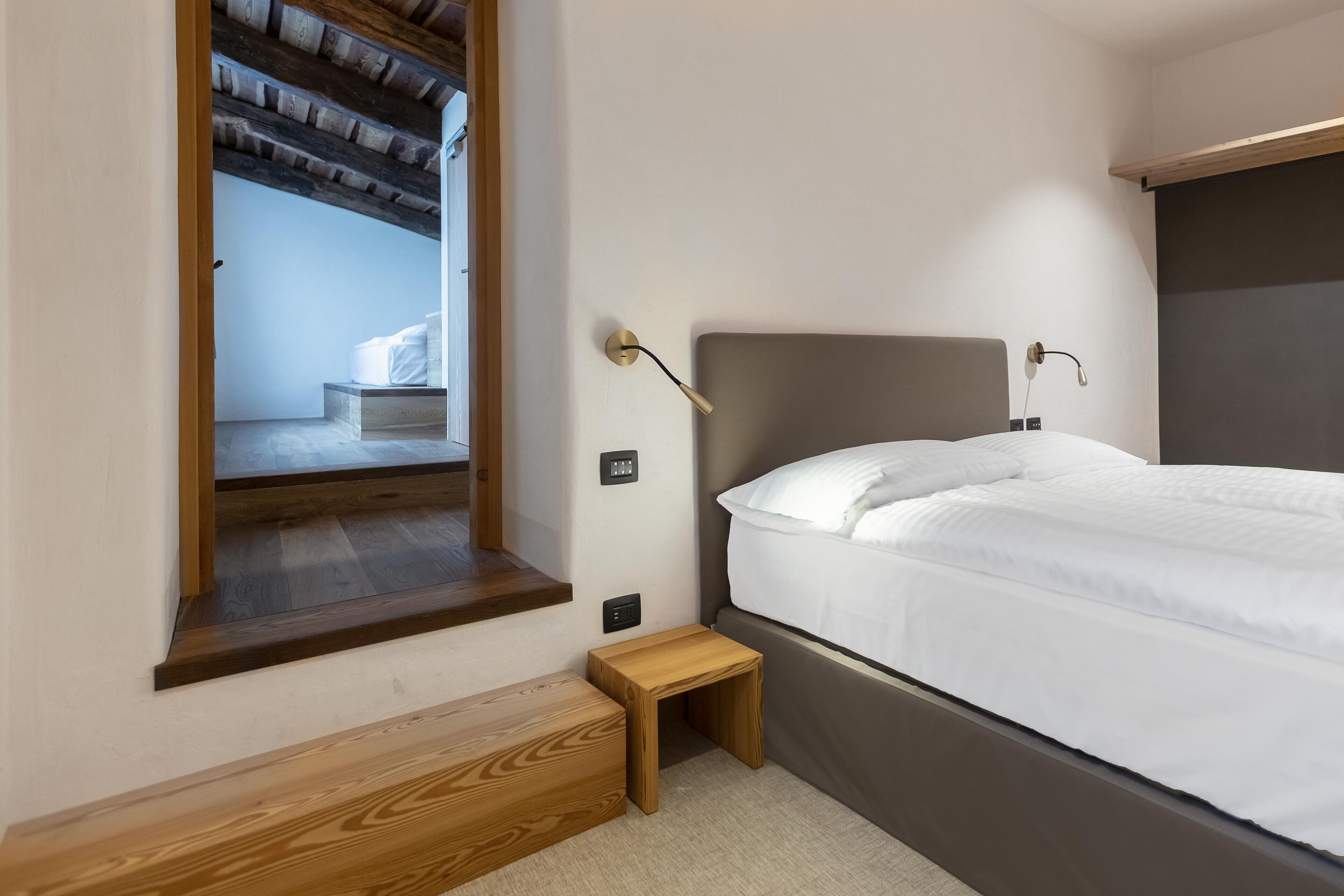 Een slaapkamer met een bed, een klein houten krukje, een nachtkastje en een raam met uitzicht op een andere kamer.