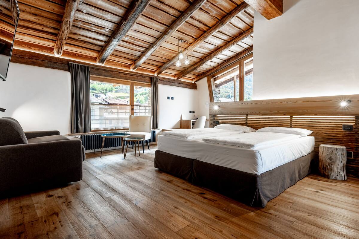Ein Schlafzimmer mit Holzboden, einem Bett, einem Sessel, einem Tisch und Blick ins Freie.