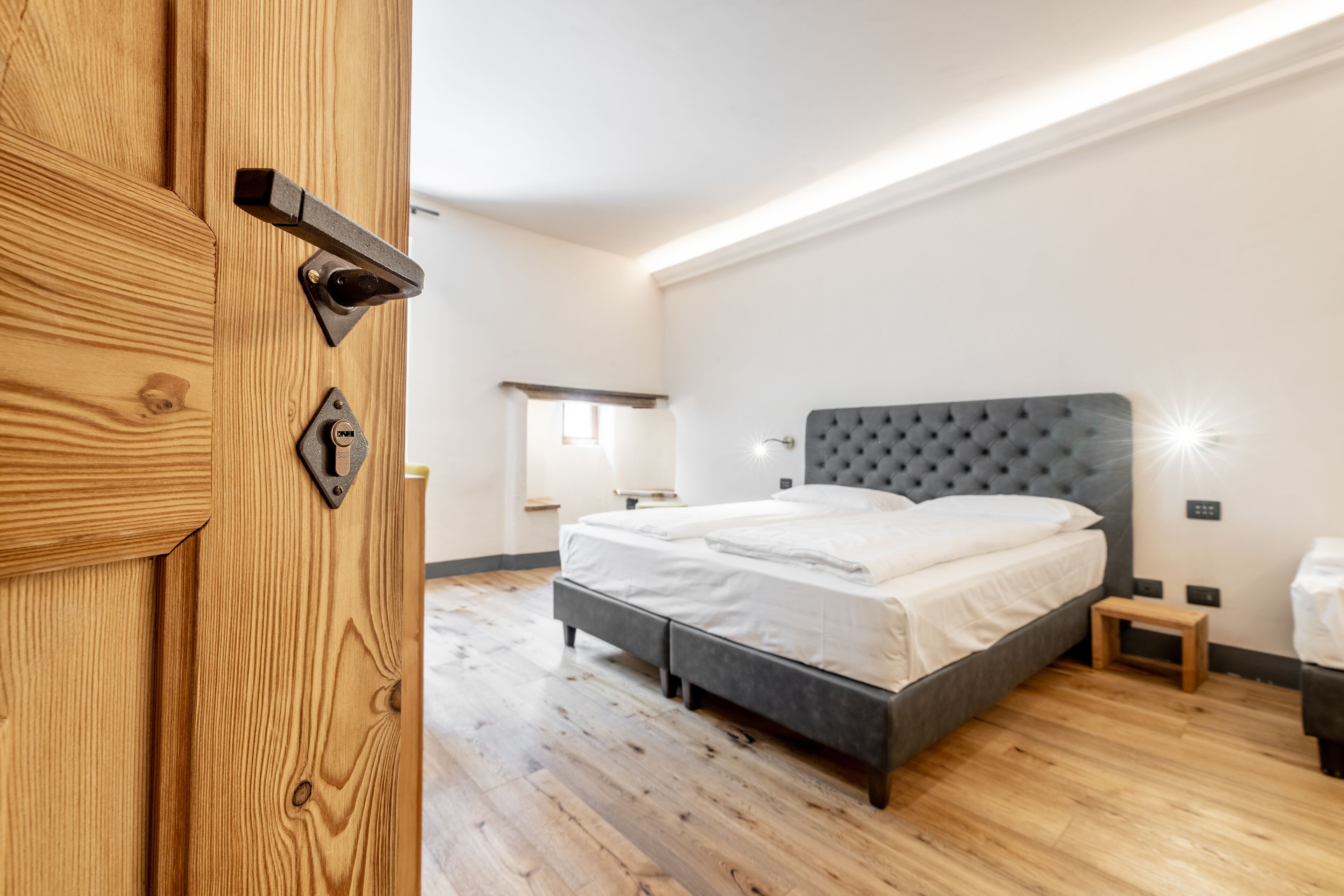 A modern bedroom with a wooden door, a bed with a grey headboard, and a small bedside table.