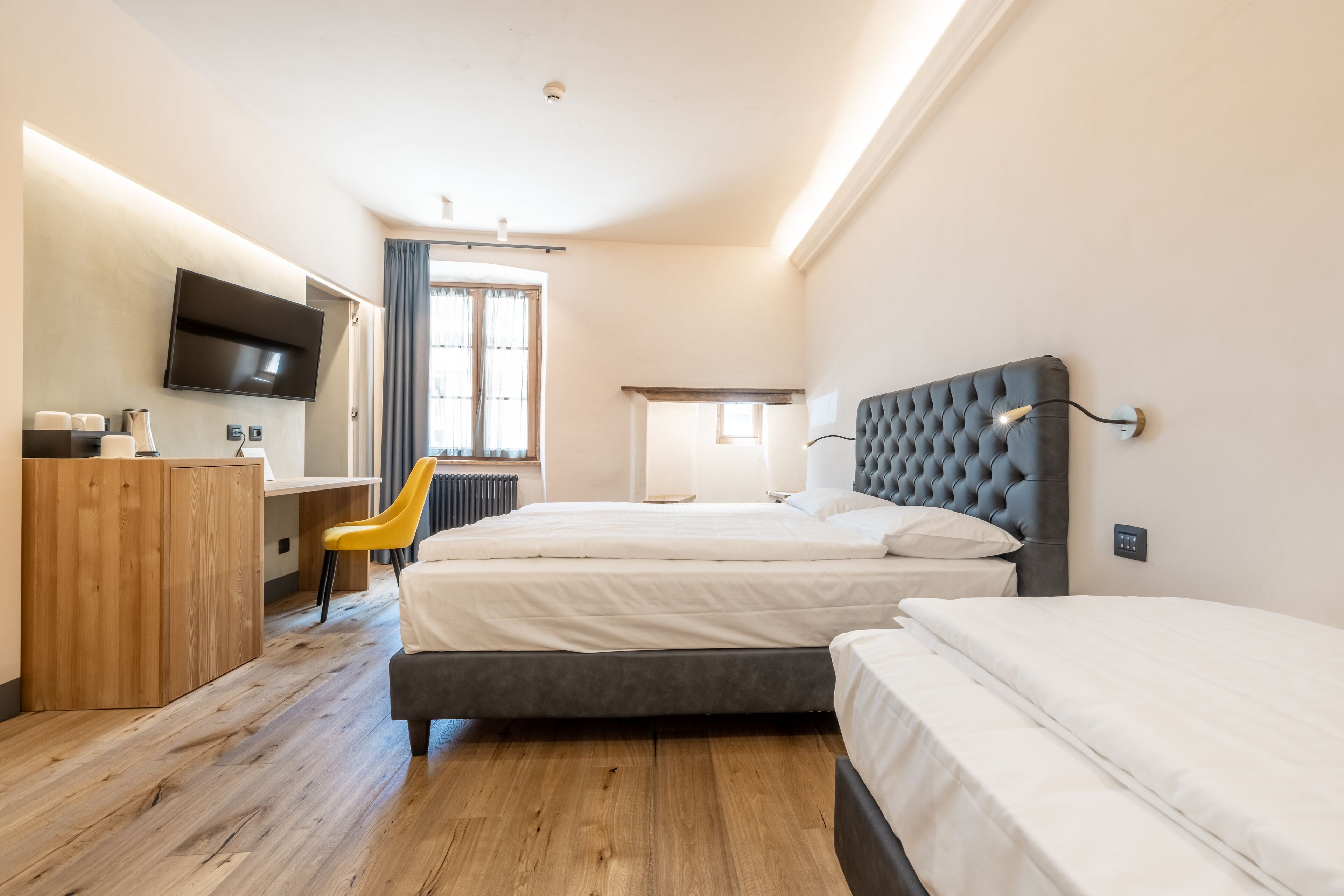 Ein Hotelzimmer mit zwei Betten, einem Schreibtisch mit Stuhl, einem an der Wand montierten Fernseher und einem Fenster mit Vorhängen.
