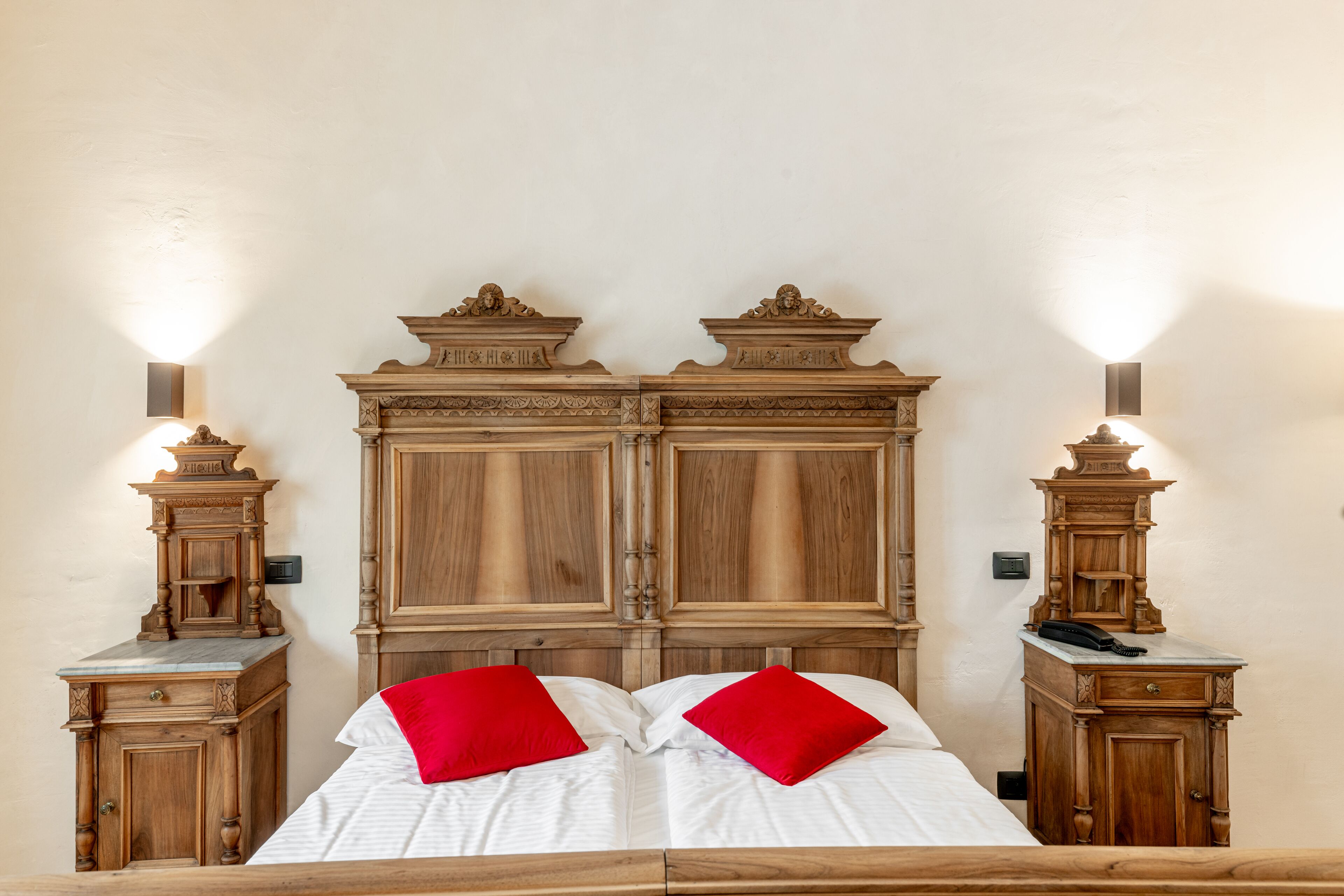 Uma cama de casal com duas almofadas vermelhas, cabeceira de madeira e mesas de cabeceira com luminárias.
