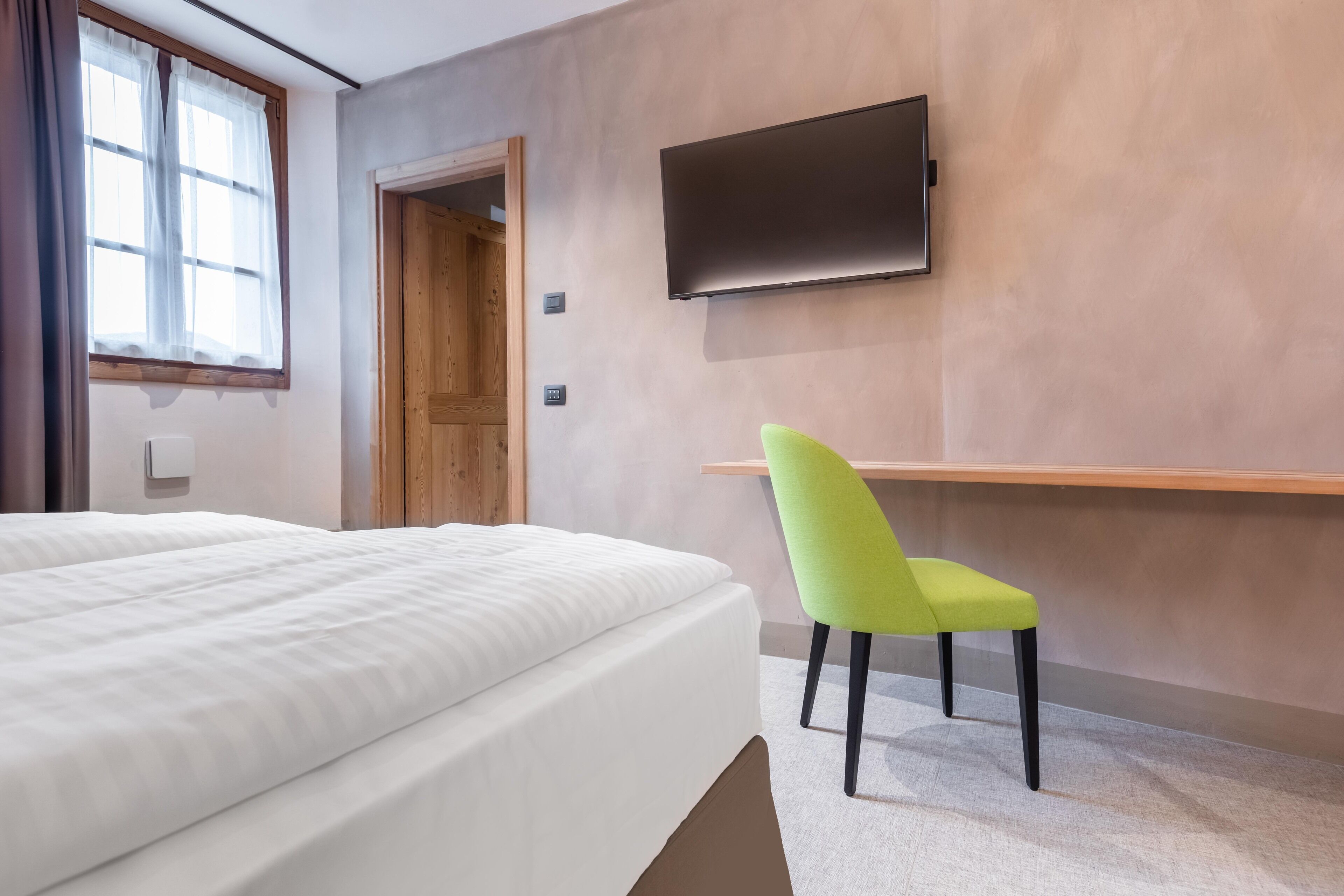Une chambre d’hôtel avec un lit, un fauteuil vert, un téléviseur fixé au mur et une porte en bois.