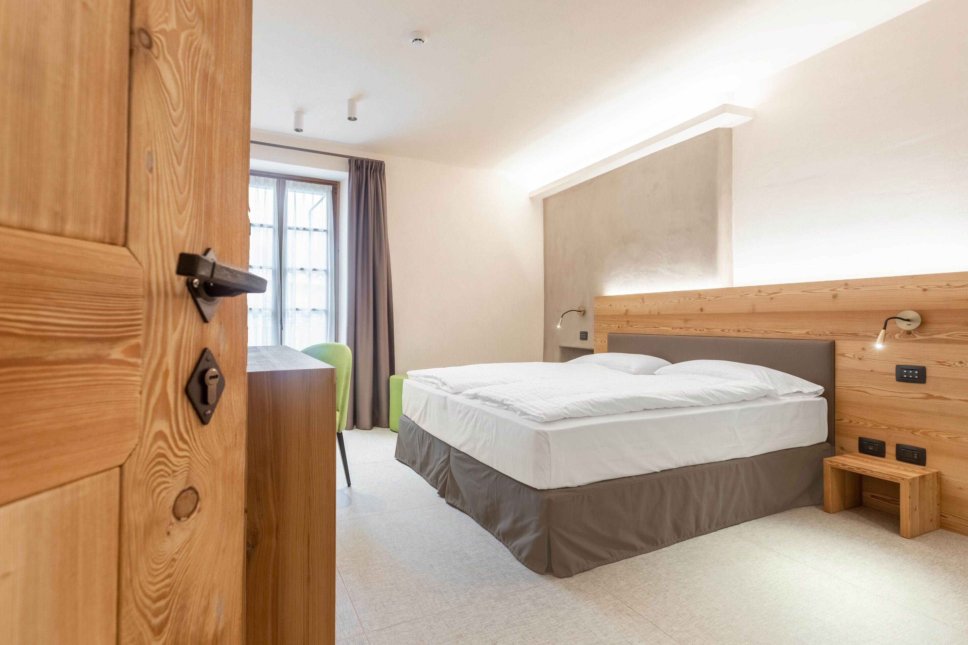 A modern hotel room with a wooden wardrobe, a bed with white linens, a bedside table, and a window with curtains.