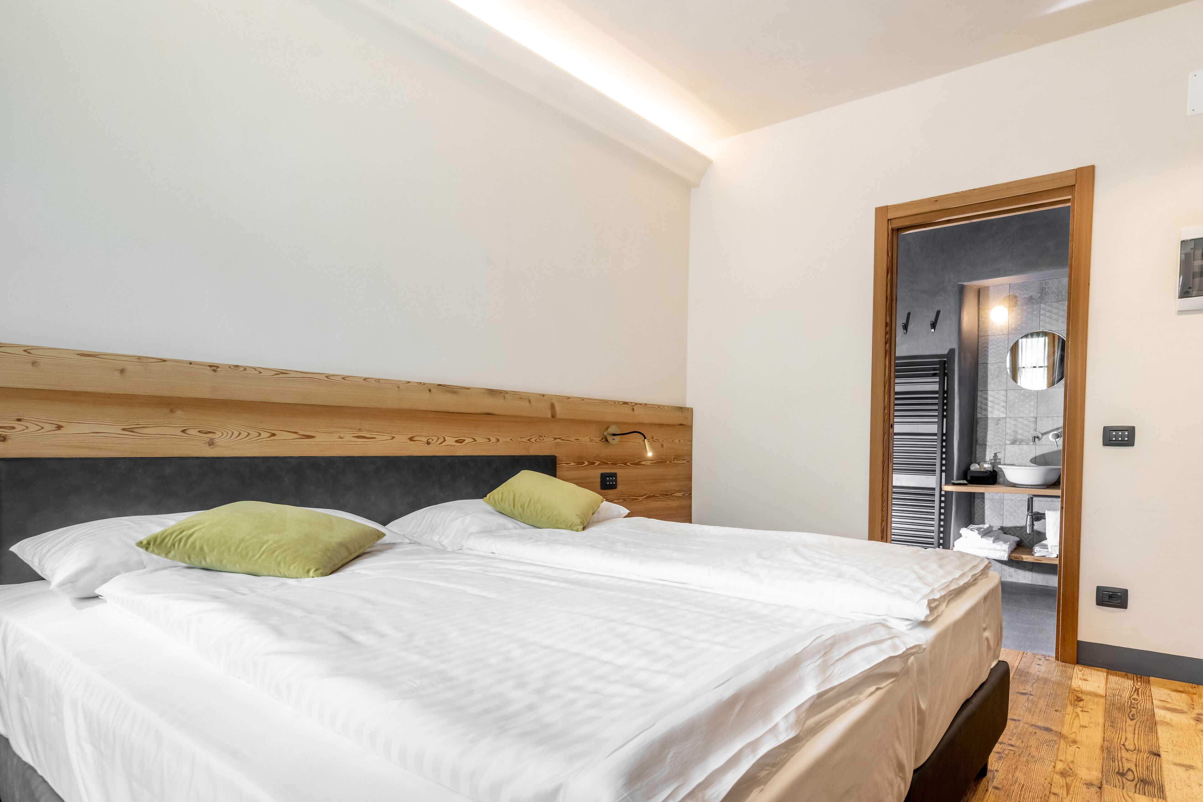 A modern bedroom with a wooden headboard, a large bed with white bedding and green pillows, a bedside table, and a view into the bathroom.